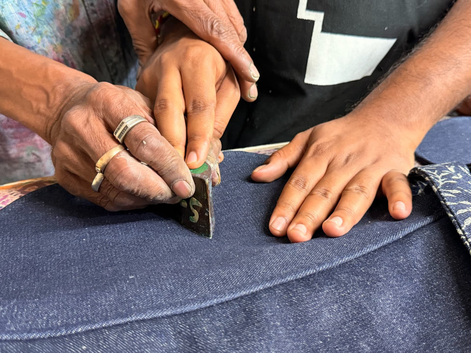 A teacher's ringed hands guide a maker's hand as they press a printing block onto denim together
