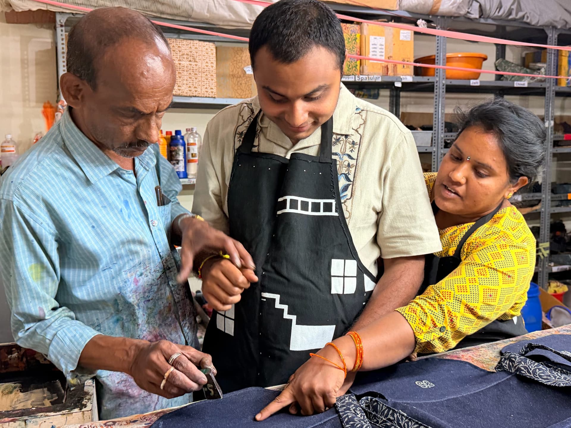 Three makers work together at the printing table, one guiding a carved block across a denim panel